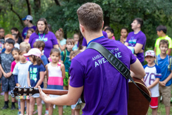 A JCamps staff member wearing a purple shirt plays the guitar for kids during Song Session.