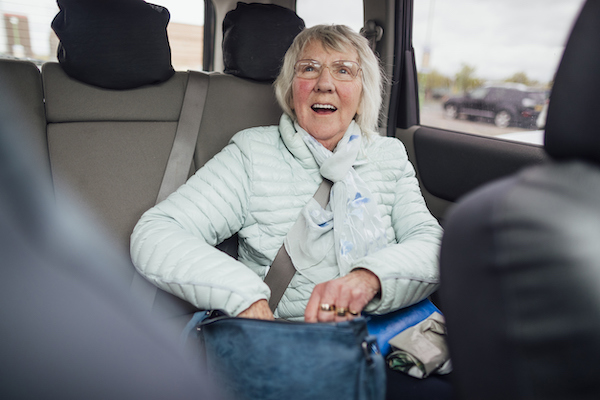 A senior woman sitting in the back of a car zipping her bag up while smiling and looking up. She is getting ready to be driven home.